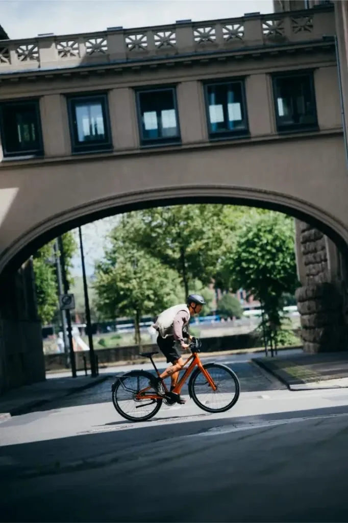 Una persona monta en una bicicleta Cannondale FlyingV naranja bajo un puente de piedra en un día soleado.