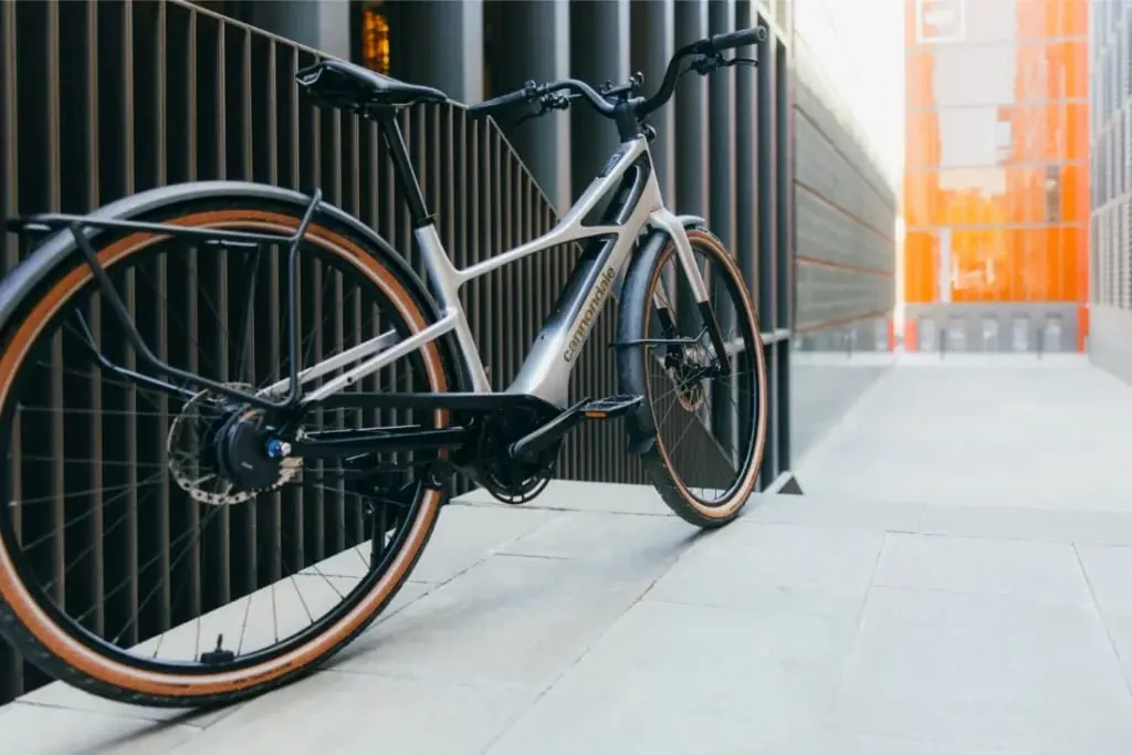 Una bicicleta eléctrica Cannondale FlyingV plateada está aparcada cerca de un moderno edificio con elementos arquitectónicos metálicos y naranjas.