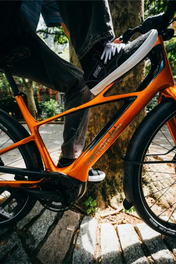 Una persona pedalea con una bicicleta Cannondale FlyingV naranja por un camino soleado y arbolado con pavimento de piedra.
