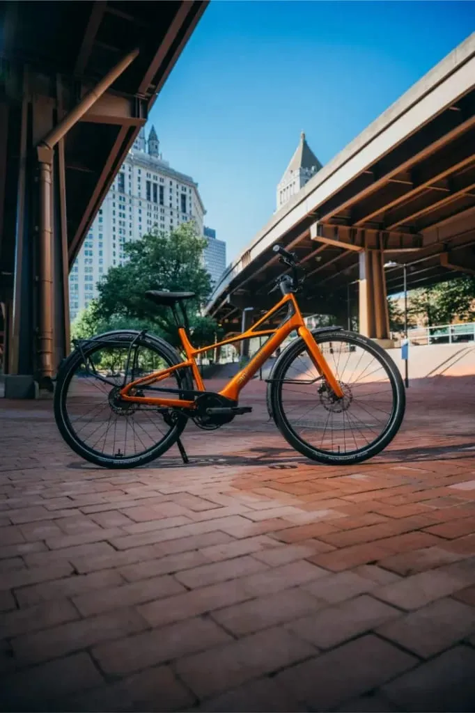 Una bicicleta Cannondale FlyingV naranja aparcada en un camino de ladrillos bajo los puentes de la ciudad, con edificios y árboles al fondo.