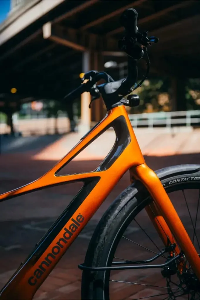Primer plano del cuadro y el manillar de una bicicleta Cannondale FlyingV naranja bajo un paso elevado, con fondo borroso.