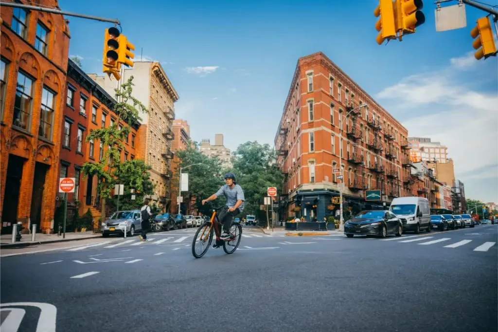 Un hombre monta en una bicicleta Cannondale FlyingV a través de una intersección de la ciudad bordeada de edificios de ladrillo rojo y coches en un día soleado.