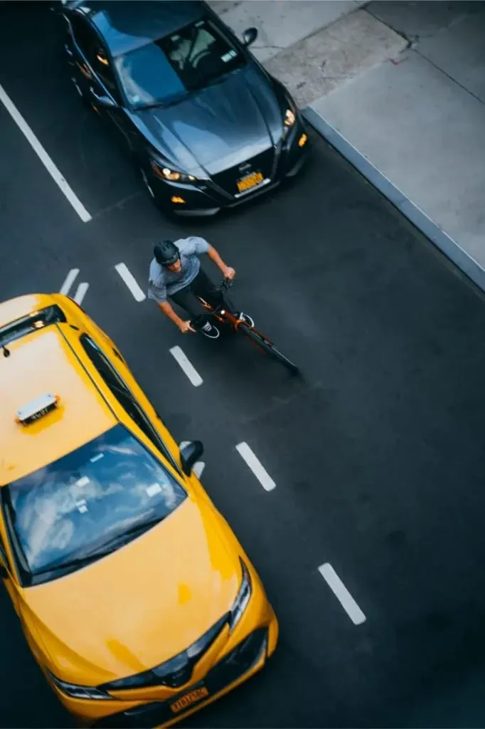 Un hombre monta en una bicicleta Cannondale FlyingV entre un taxi amarillo y un coche gris en una calle de la ciudad, vista desde arriba.