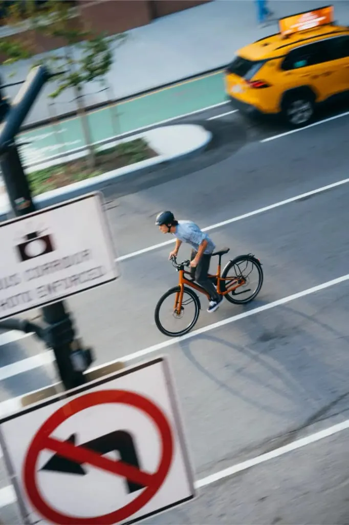 Una persona monta en una bicicleta Cannondale FlyingV naranja en una calle de la ciudad junto a un taxi, con señales de tráfico en primer plano.