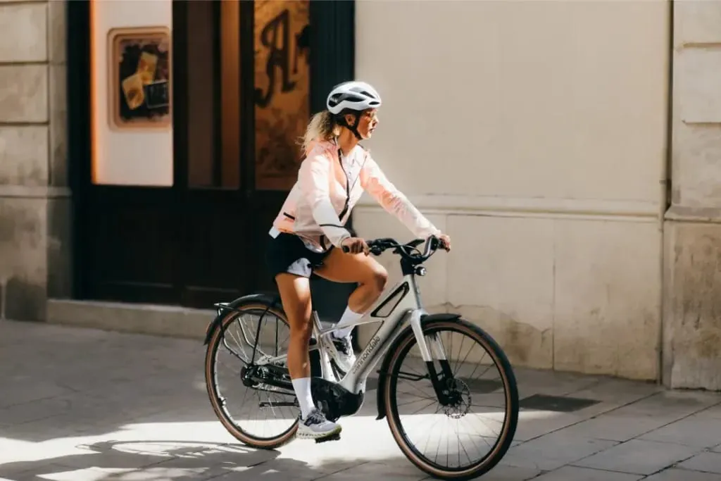Mujer con casco y chaqueta montando en bicicleta por una calle soleada de la ciudad.