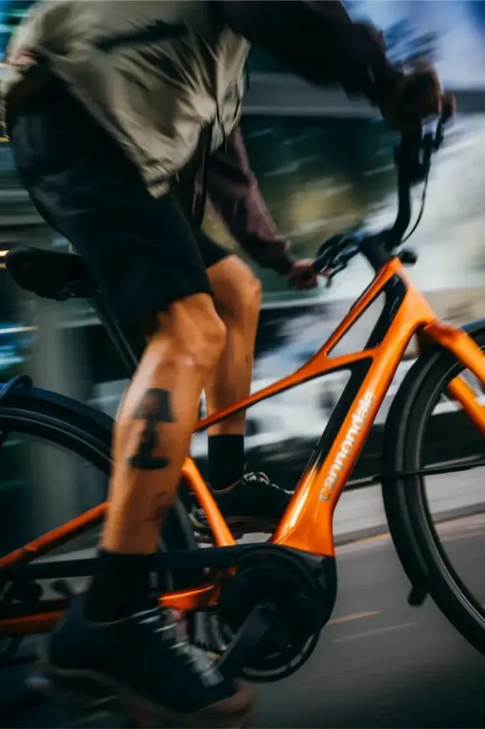 Persona montando una bicicleta Cannondale FlyingV de color naranja, mostrada de lado con un fondo borroso, enfocado en las piernas y la bicicleta.