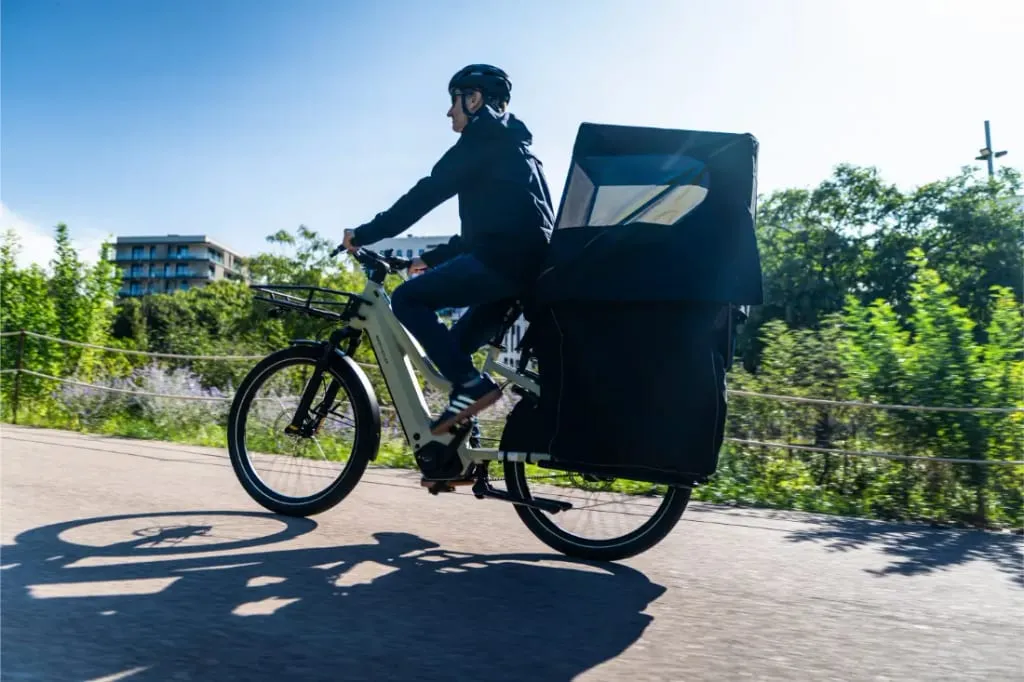 Una persona monta en una bicicleta de carga Riese & Müller Multicharger3 con un gran compartimento cubierto en un día soleado, árboles y edificios al fondo.
