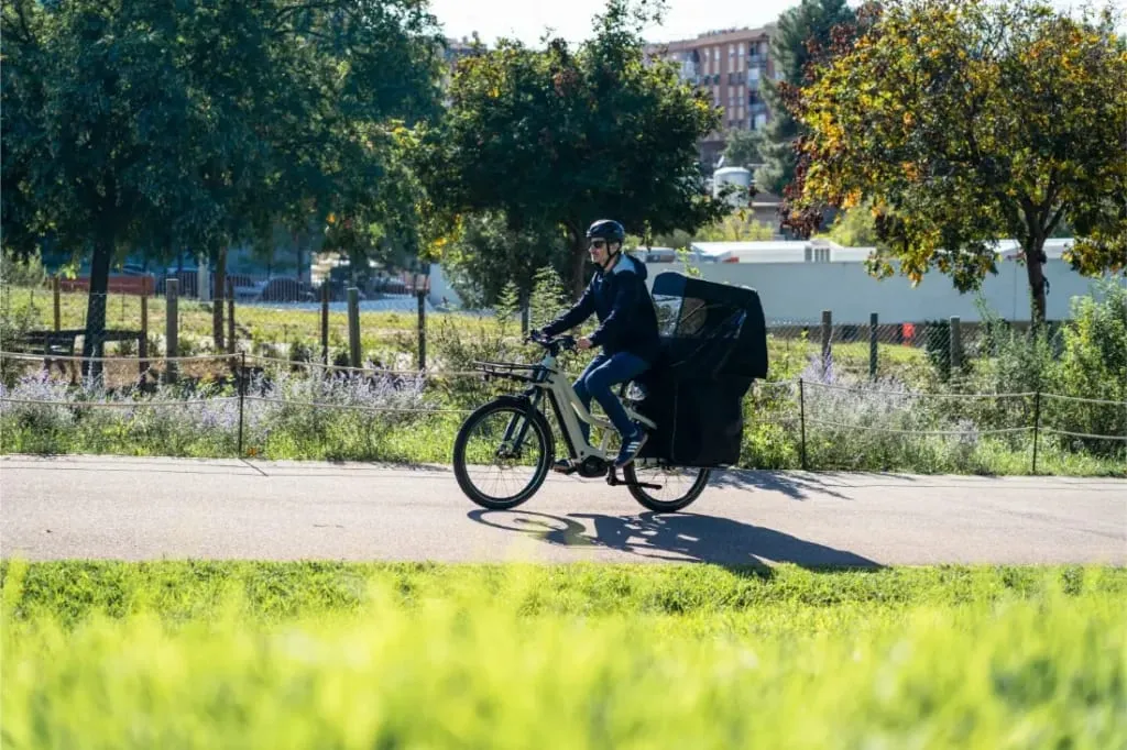 Persona montando en una e-bike Riese & Müller Multicharger3 Mixte Vario con una bolsa de carga grande por el sendero de un parque, rodeado de vegetación.