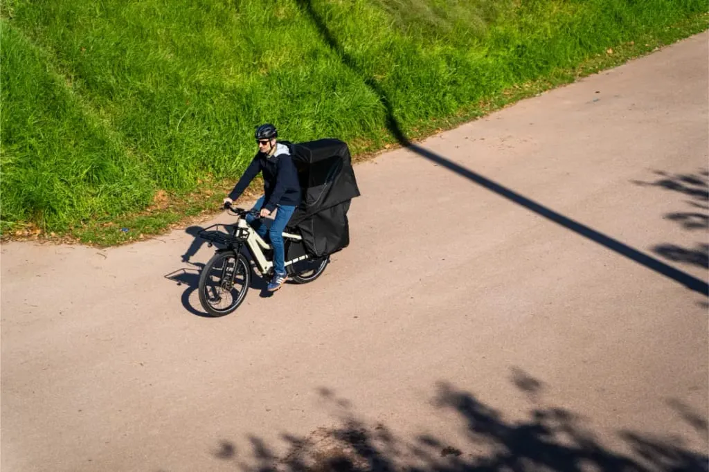 Una persona conduce una bicicleta eléctrica de carga Riese & Müller Multicharger3 Mixte Vario con una caja de entrega negra por una carretera soleada junto a hierba verde.