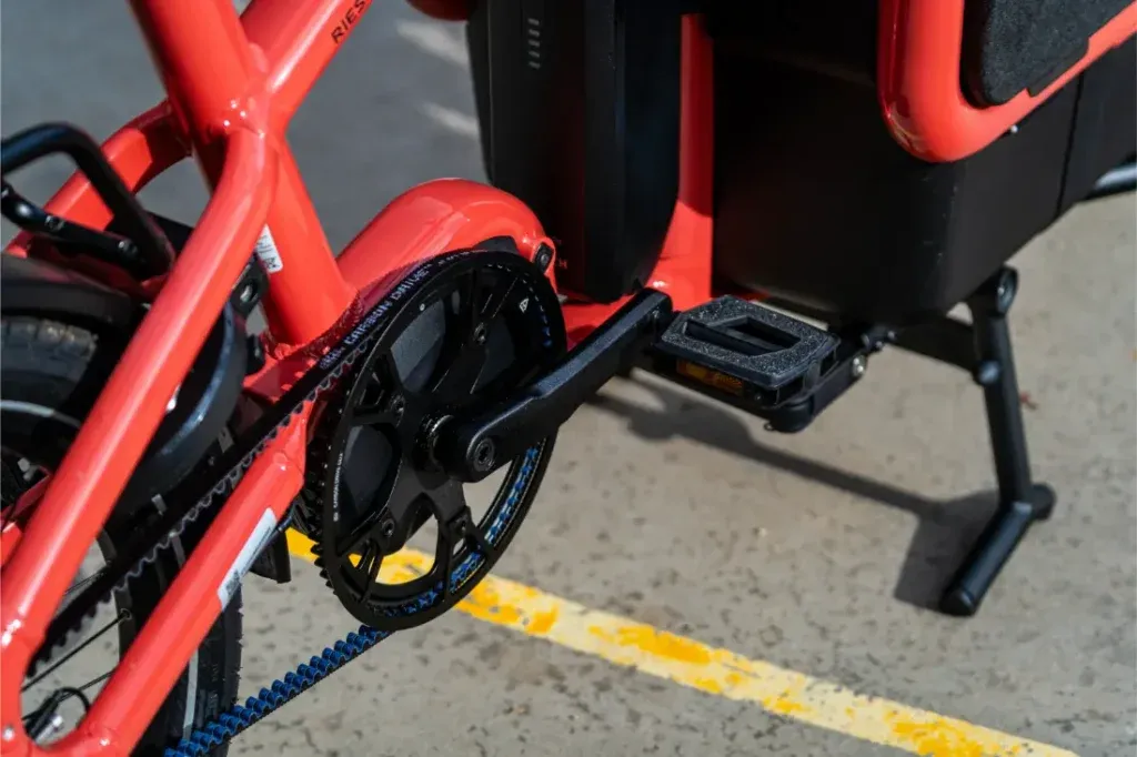 Primer plano del pedal, la biela y el plato de una bicicleta Riese Müller roja -que muestra su tamaño compacto- aparcada en una superficie de hormigón con líneas amarillas.