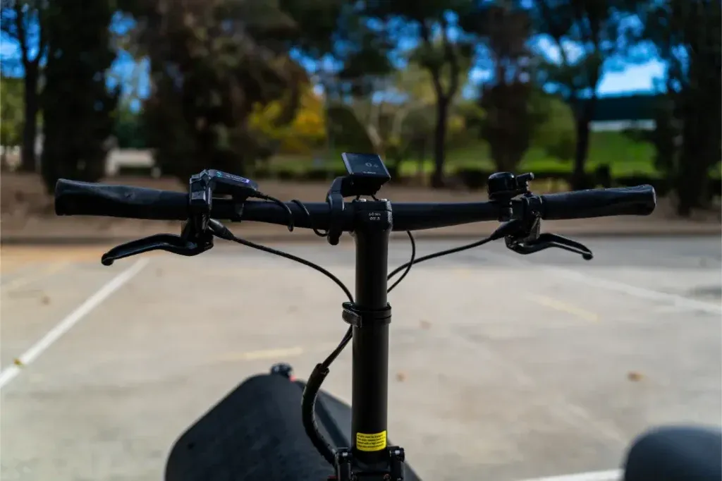 Primer plano del manillar de una bicicleta compacta Riese Müller en un aparcamiento vacío, enmarcado por árboles al fondo.