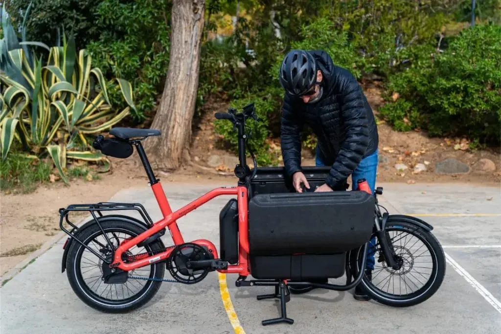 Una persona con casco ajusta el compartimento portaobjetos de una bicicleta eléctrica de carga Riese Müller Carrie2 roja, conocida por su tamaño compacto, aparcada al aire libre.