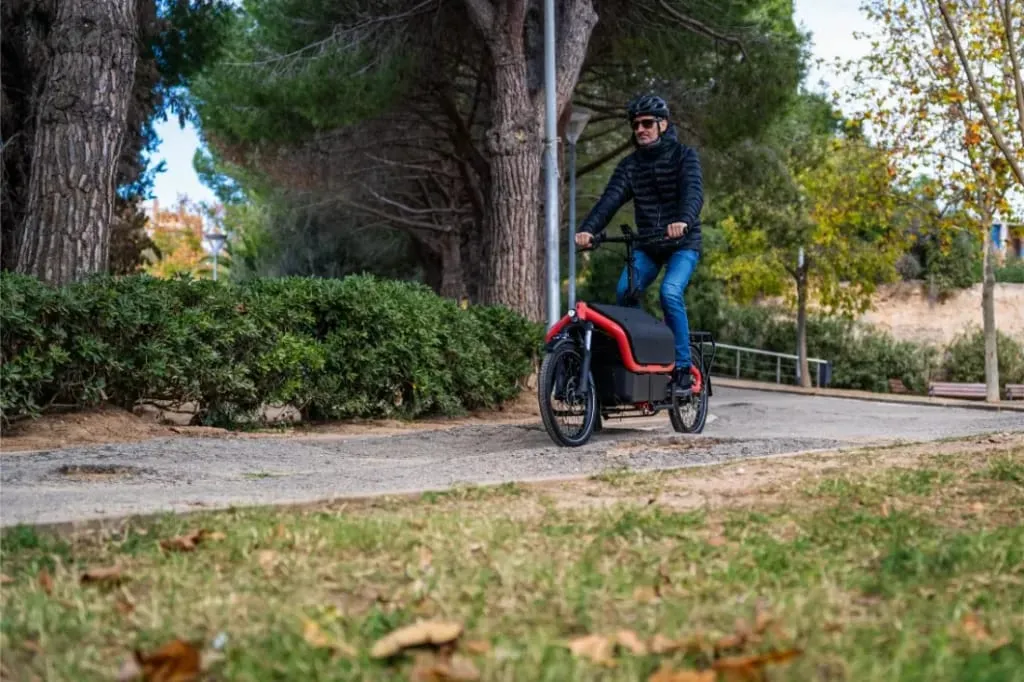Una persona con casco conduce una bicicleta eléctrica Riese Müller roja por un camino pavimentado a través de un parque con árboles.