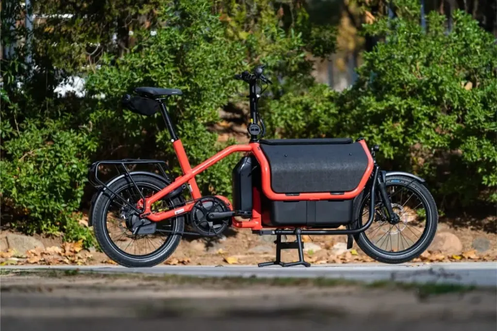 Una bicicleta de carga eléctrica automática Riese Müller Carrie2 roja con ruedas negras está aparcada en un camino pavimentado delante de arbustos verdes, mostrando su tamaño compacto y su diseño moderno.