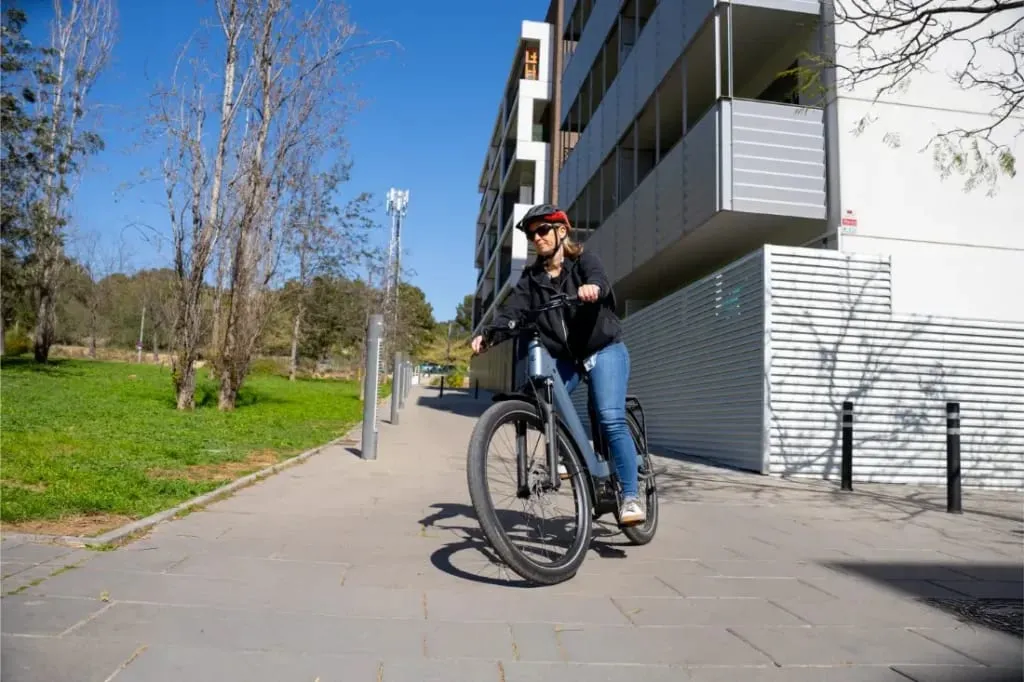 Mujer montando en una bicicleta Riese & Müller Nevo5 en una acera cerca de edificios modernos y una zona de parque con césped, disfrutando tanto del lujo accesible como de la silenciosa tecnología CORE en un día soleado.