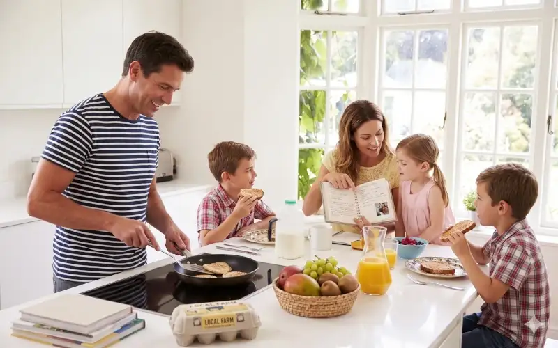 Dad cooks as mom reads to daughter; son eats in a bright, happy kitchen.