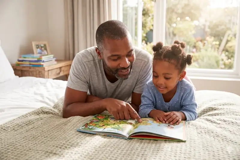 Dad and happy daughter reading a storybook together in bed.