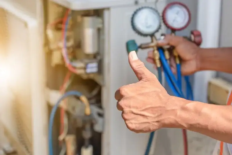 HVAC technician gives thumbs-up while holding manifold gauges, servicing an AC unit.