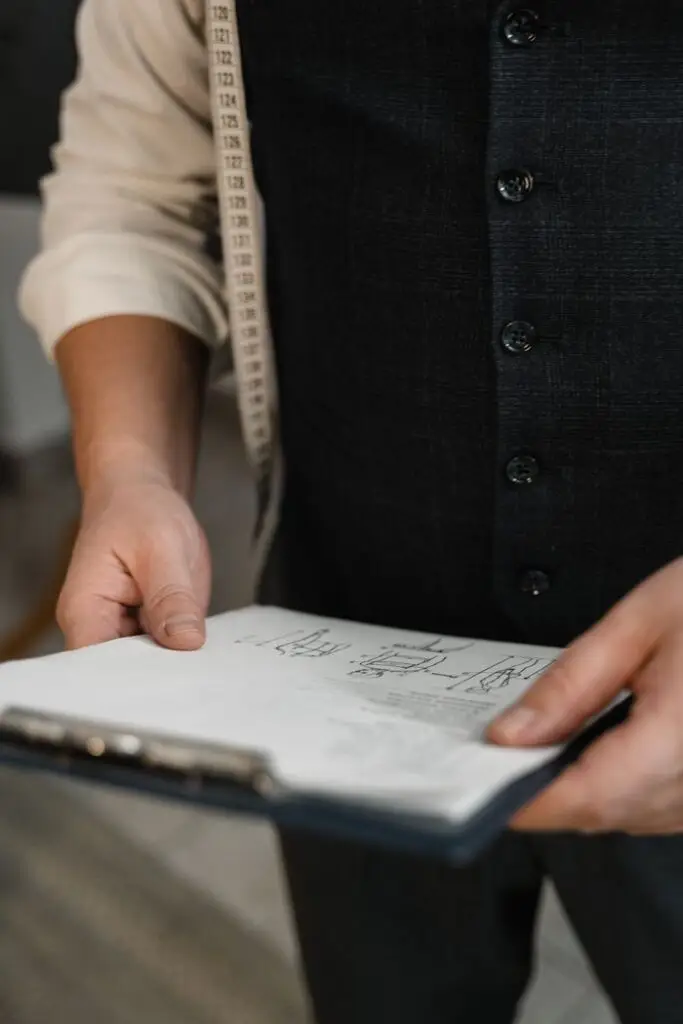 A tailor with measuring tape holds a clipboard with sketches, highlighting precision work.