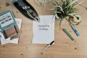 A wooden desk with a wrinkled paper labeled “Marketing Strategy,” a black iron, a potted plant, an open marker, tape, and two marketing books scattered around.