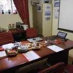 A conference table setup with snacks, papers, and a laptop; coffee station and window with red curtains in the background.
