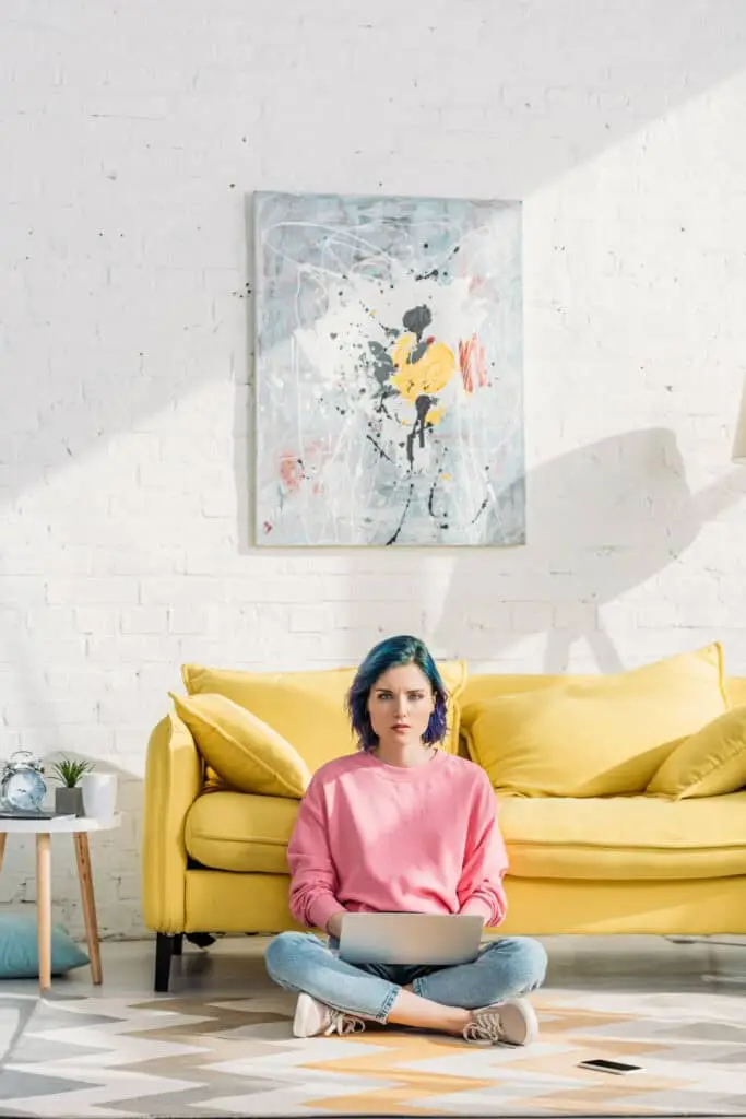 Fabre Occitanie - Jeune femme aux cheveux bleus assise en tailleur sur un tapis, utilisant un ordinateur portable devant un canapé jaune, avec des œuvres de Fabre Occitanie au mur.