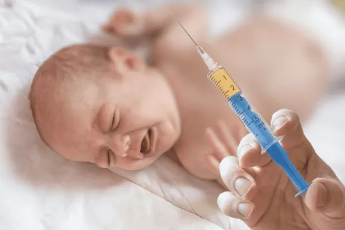 En hand håller en K-vitaminspruta framför ett gråtande spädbarn