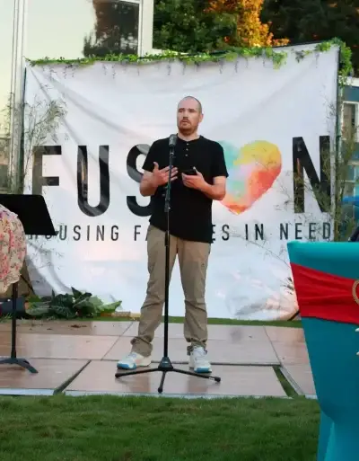 Man speaks at microphone on outdoor stage. "FUSION HOUSING FAMILIES IN NEED" banner behind.