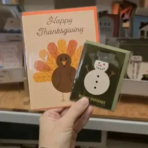 Hand holding a Happy Thanksgiving turkey card and a Happy Holidays snowman card.