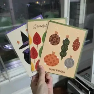 Hand holding three colorful seasonal cards: Halloween witch, autumn leaves, and holiday ornaments.