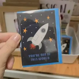 Hand holding a card with a white rocket, stars, and "YOU'RE OUT OF THIS WORLD" text.