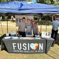 Three smiling FUSION Housing volunteers with a heart logo at an outdoor event.