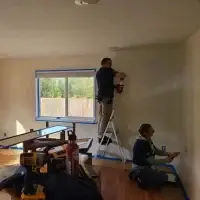 Man on ladder spraying, woman kneeling with roller, both painting a room with blue tape.
