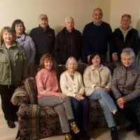 Ten smiling adults: five standing behind five seated on a patterned sofa, indoors.