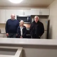 Three smiling older adults stand in a modern kitchen with white cabinets and new appliances.