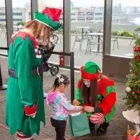 Child receives gift from two festive elves next to a Christmas tree.