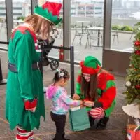 Two Christmas elves give a gift from a green bag to a small child.
