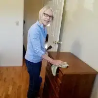Smiling senior woman in glasses dusting a brown wooden dresser with green cloth.