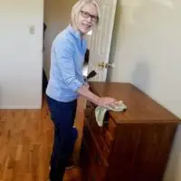 Smiling older woman with glasses dusting a wooden dresser with a green cloth.