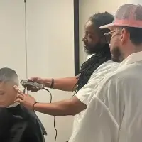 Barber with dreadlocks trims a child's hair as an apprentice observes.