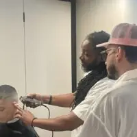 A barber with dreadlocks gives a boy a fade haircut, observed by another man.