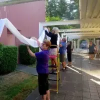 Community members hang white fabric decorations on an outdoor pergola walkway.
