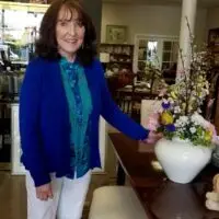 Smiling older woman in blue cardigan, teal shirt, by colorful flowers in a white vase.