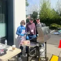 Three smiling adults barbecue outdoors. Woman holds tongs, man flashes peace sign.