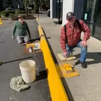 Two older men kneel, painting a curb bright yellow on a sunny day.