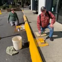 Two men kneeling, painting a long curb bright yellow on a sunny day.