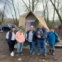 Five smiling, bundled people stand before a tent with an American flag.