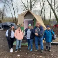 Five smiling, warmly dressed people stand before a makeshift shelter displaying an American flag.