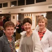 Three smiling older women at a bustling indoor social event.