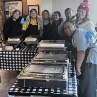 Seven smiling women and girls behind a black-and-white checkered buffet table with chafing dishes.